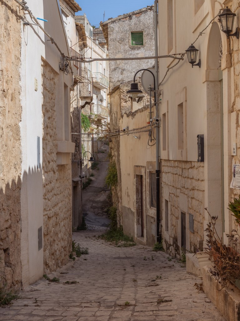 Narrow descending lane with pale façades, rough stone walls, overhead wires, black lamps, weeds, and a single green shutter.
