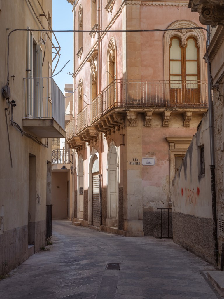 Shaded alley beside a faded pink Baroque building, with ornate balconies, overhead cables, graffiti, and an empty street.