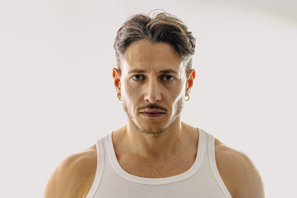 Close frontal portrait of a man in a white vest looking directly at the camera.