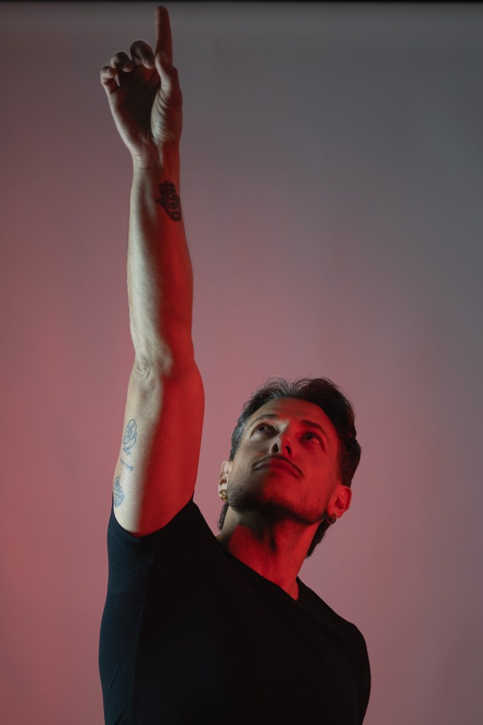 Man in a black shirt raising one arm and pointing upward under red light.