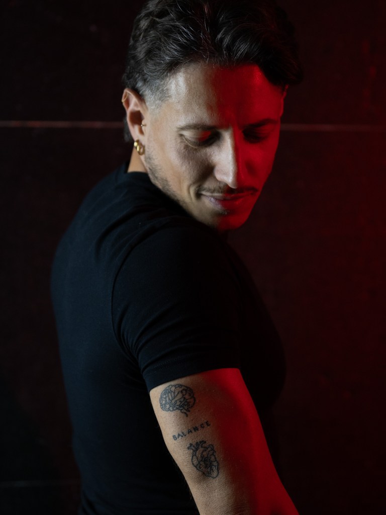 Man looking down toward tattoos on his arm, lit by red and neutral light.