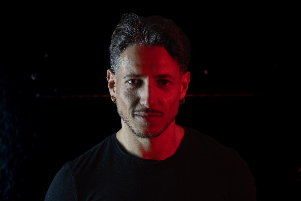 Frontal portrait of a man in a black shirt, half illuminated by red light against a dark background.