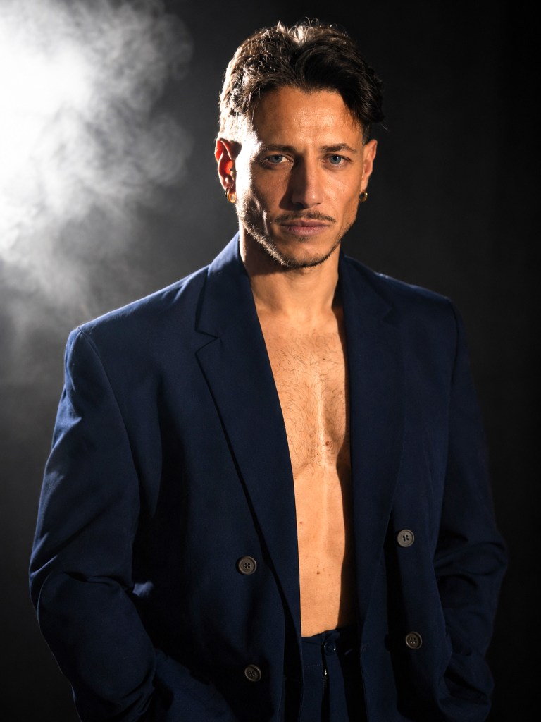 Man wearing an open dark jacket, standing before smoke and a bright backlight.