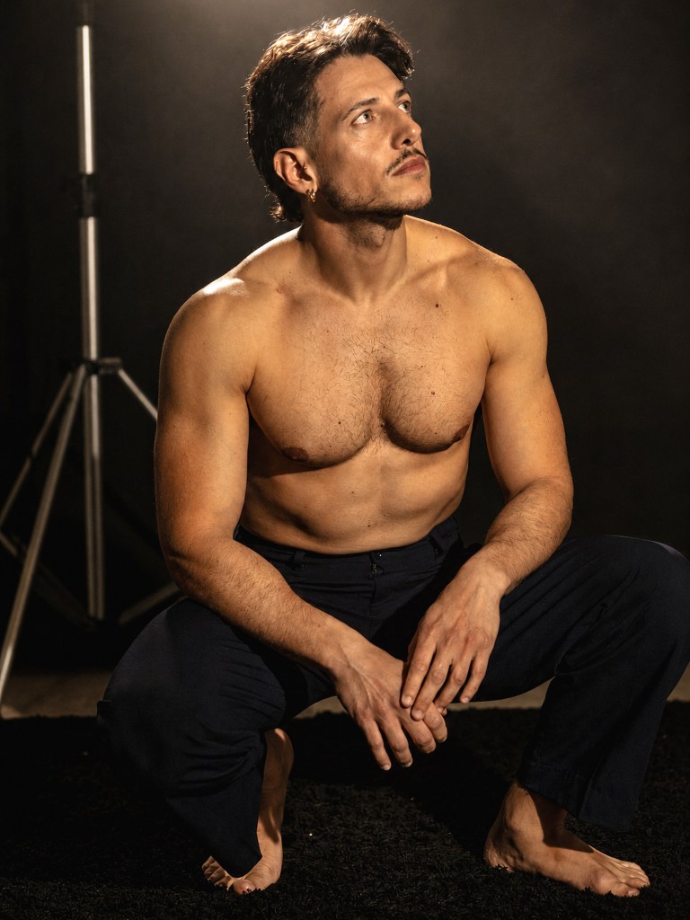 Shirtless man seated barefoot in dark trousers, looking upward under warm studio light.
